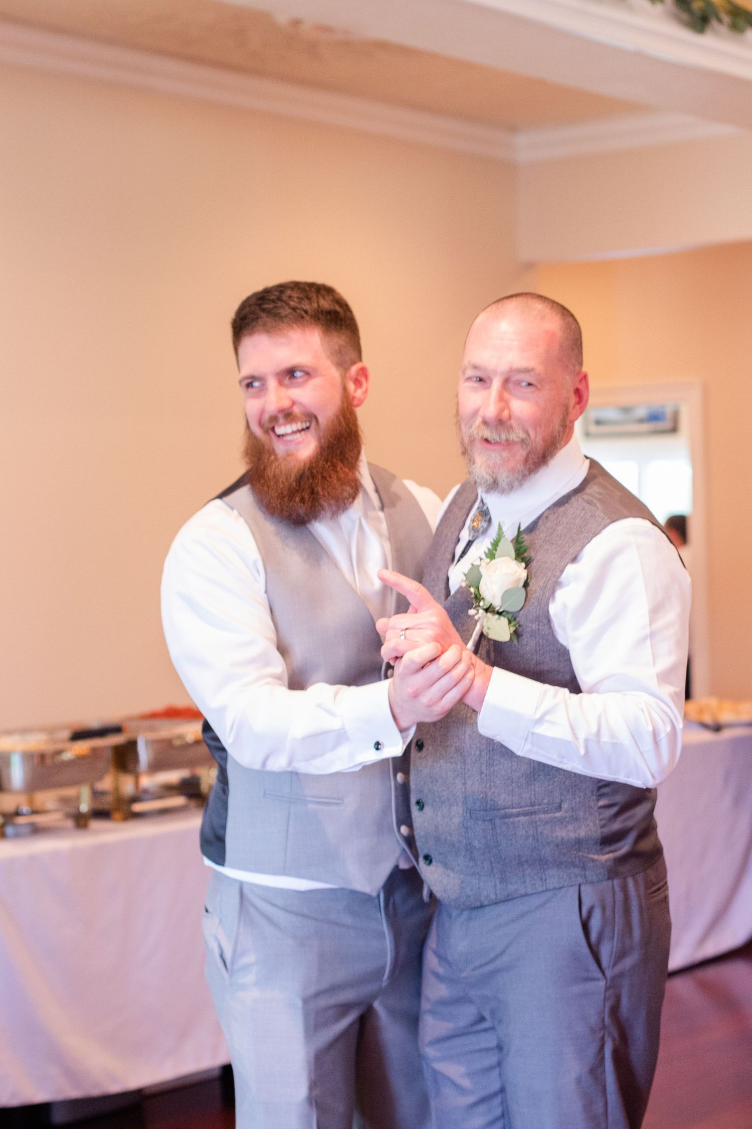 Groomsmen in matching grey vests share laughs and embraces during wedding reception.
