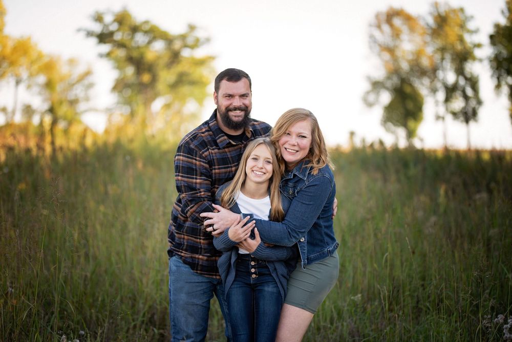 Family of three smiling together in a golden field at sunset, parents hugging their daughter.