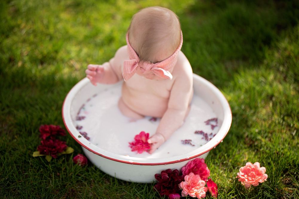 Baby sitting in a milk bath bowl with flower petals on a green grass lawn outdoors.