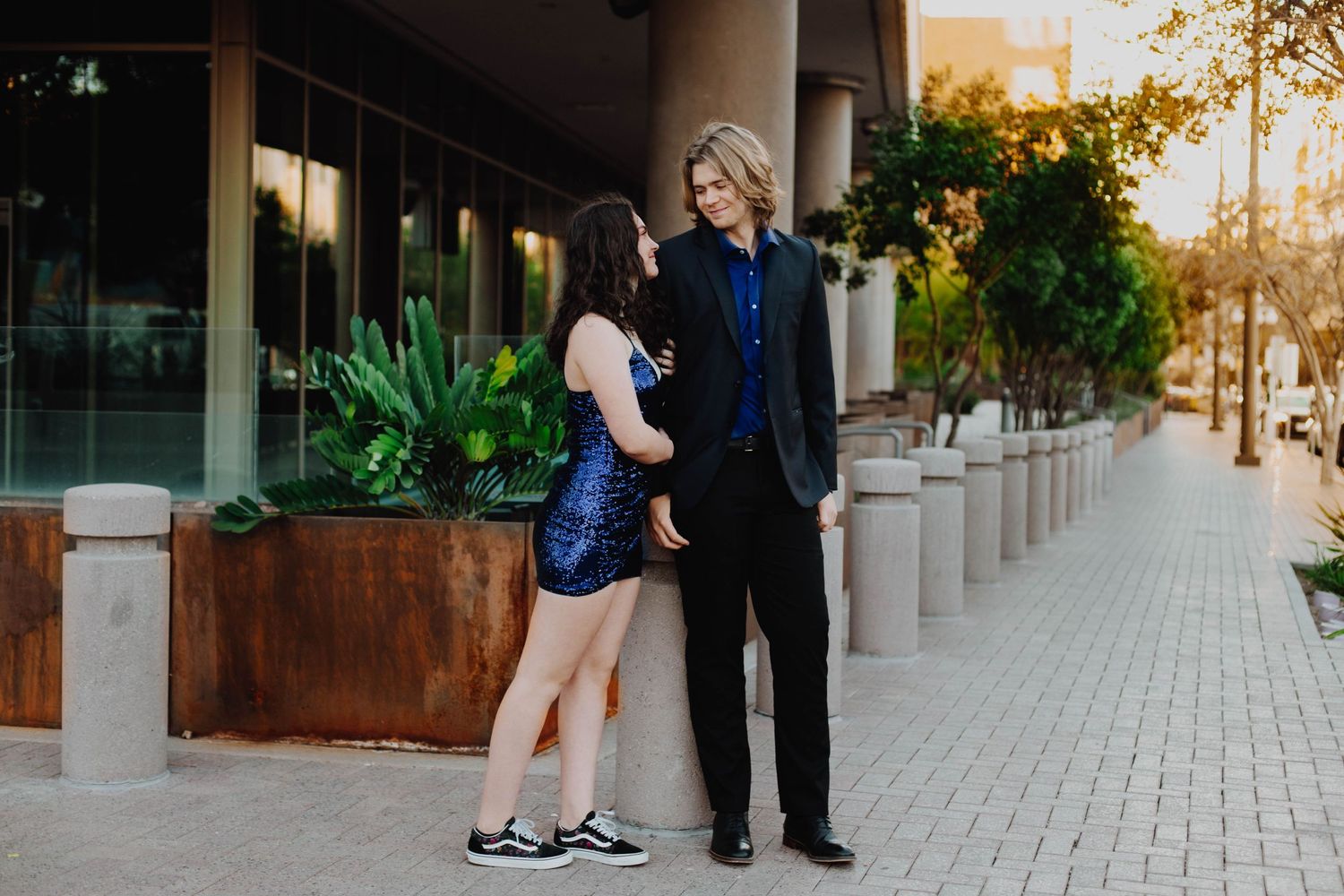 Couple in contrasting casual and formal wear standing together on an urban sidewalk at sunset.