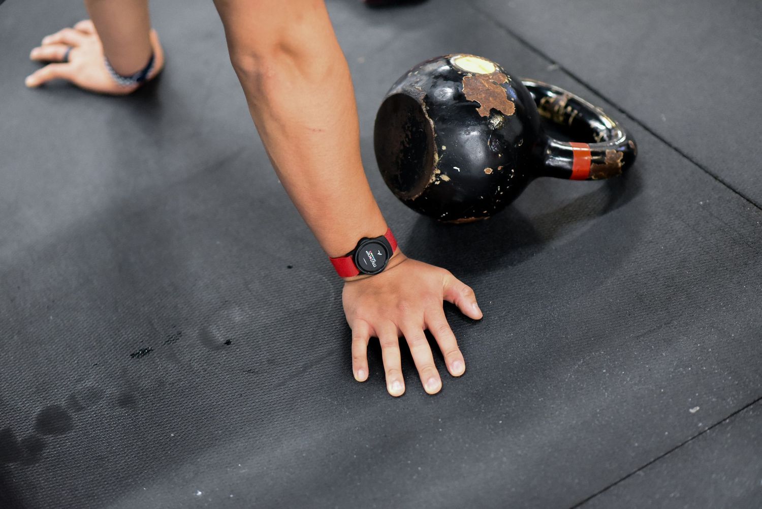Close-up of hand next to black kettlebell on dark gym floor with fitness watch visible.