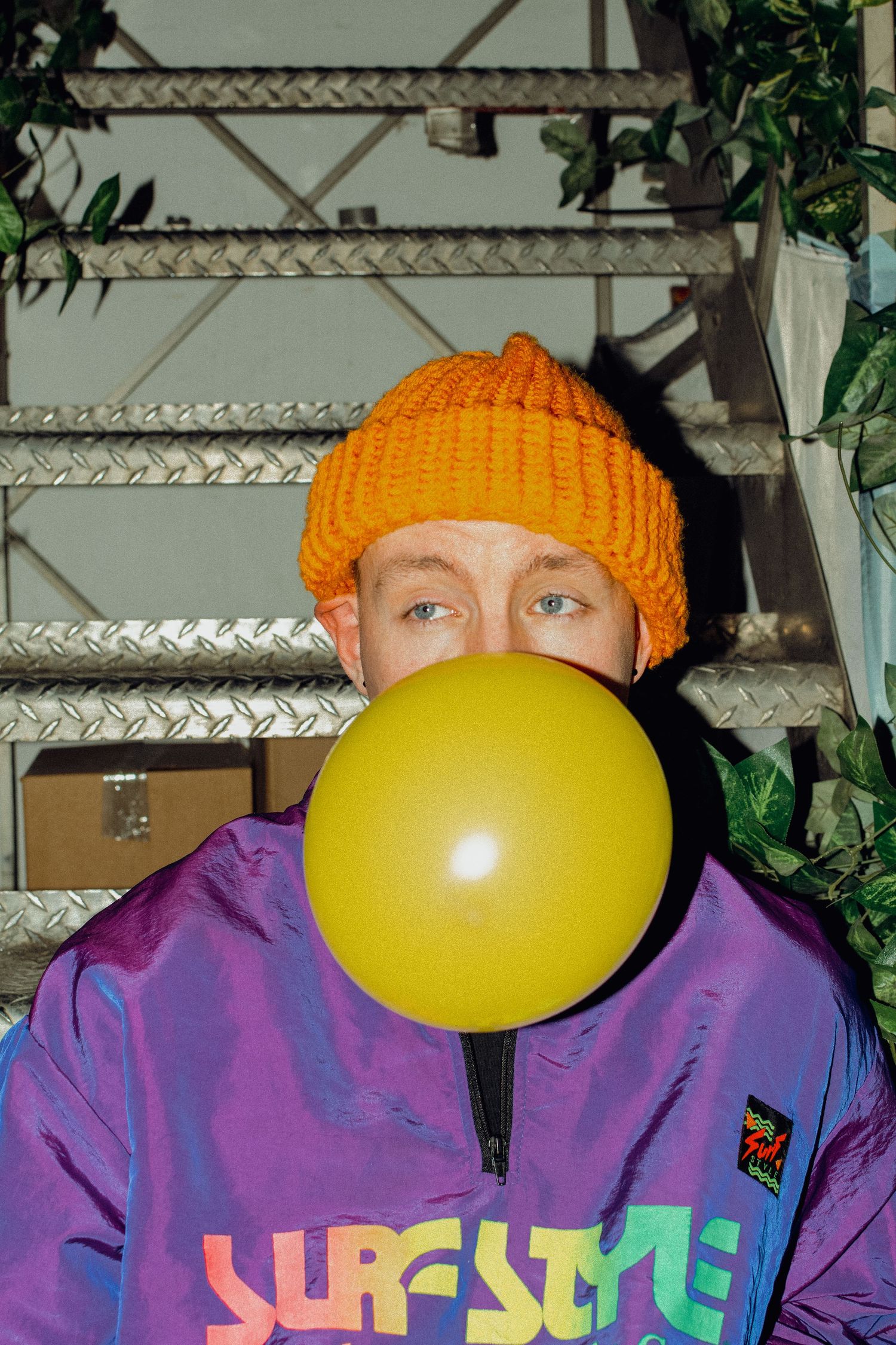 A playful shot of someone in a purple jacket and orange beanie blowing a yellow balloon.