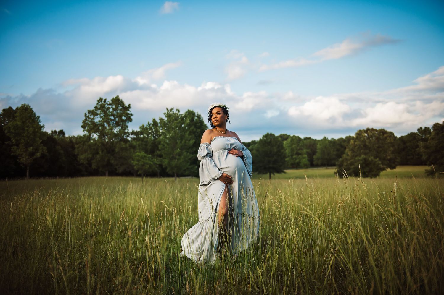 Ethereal maternity portrait in flowing white dress amid tall grass at sunset.