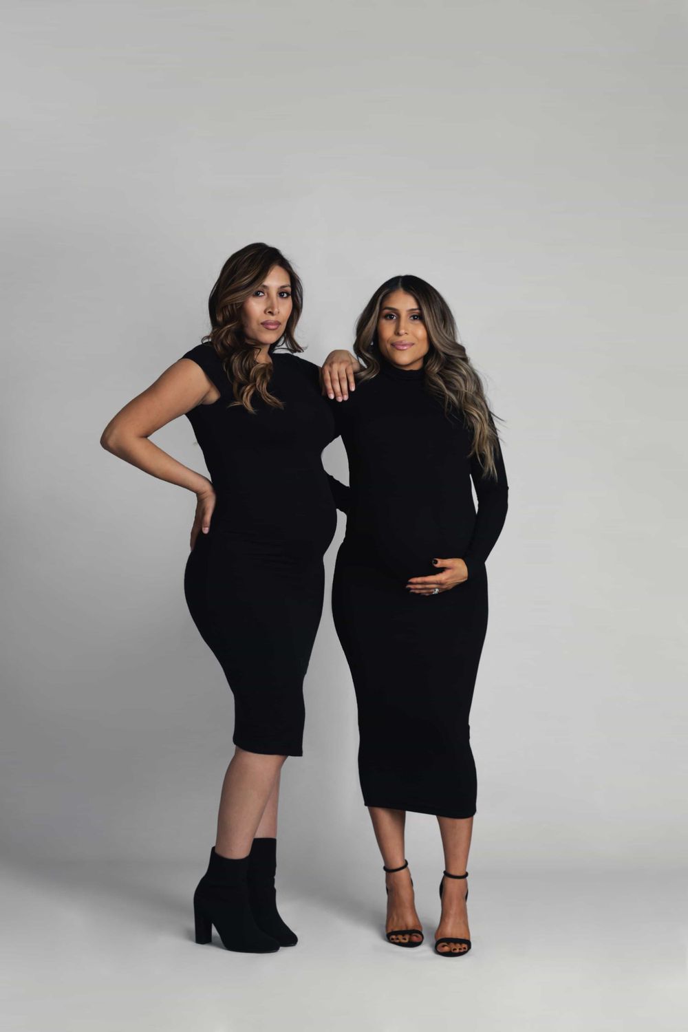 Two women in matching black midi dresses pose together, one visibly pregnant, against a neutral gray background.
