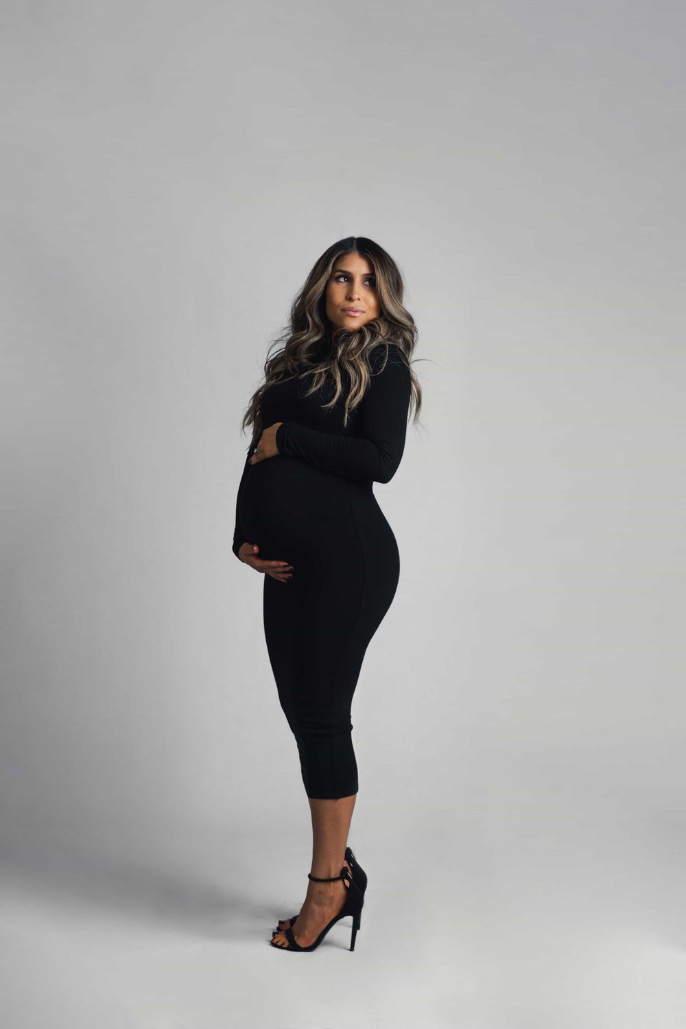 Pregnant woman in fitted black long-sleeve midi dress and heels poses sideways showing her baby bump.
