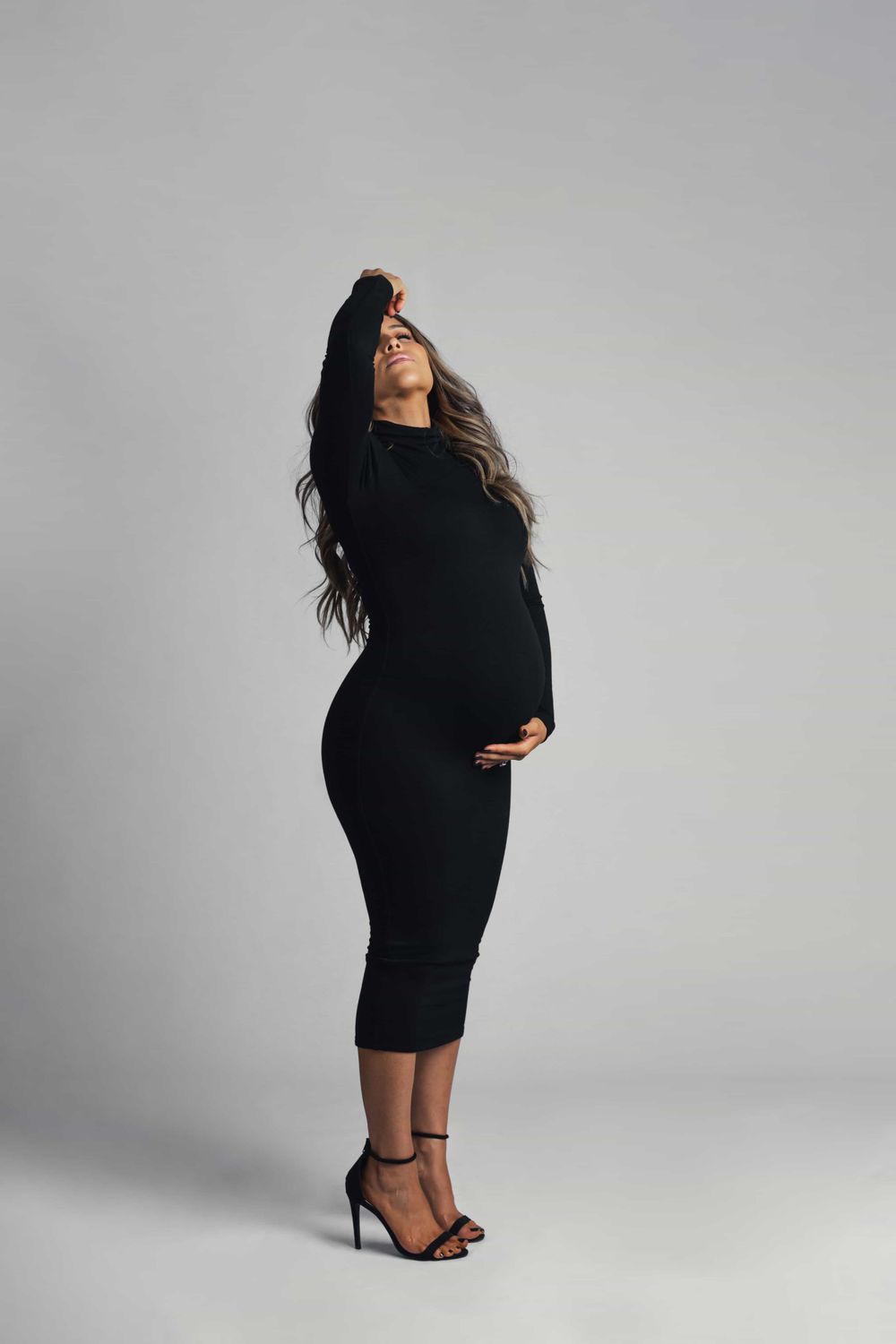 Pregnant woman in black bodycon dress and strappy heels poses dramatically with hand raised against gray backdrop.
