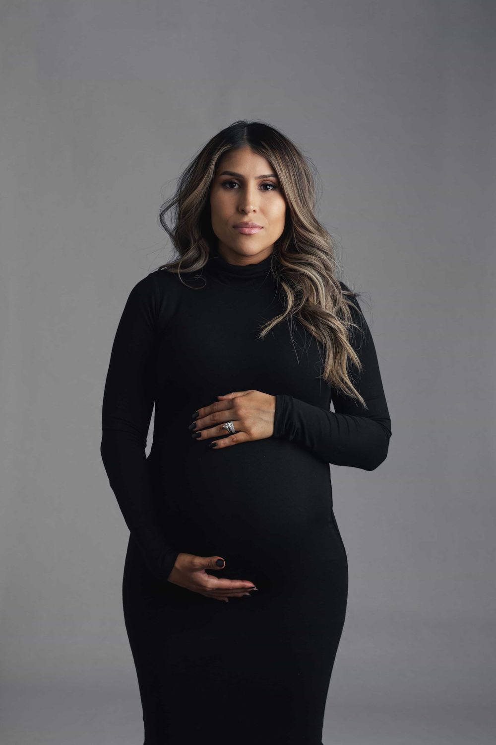 Pregnant woman in fitted black turtleneck dress cradles baby bump while gazing at camera in studio.