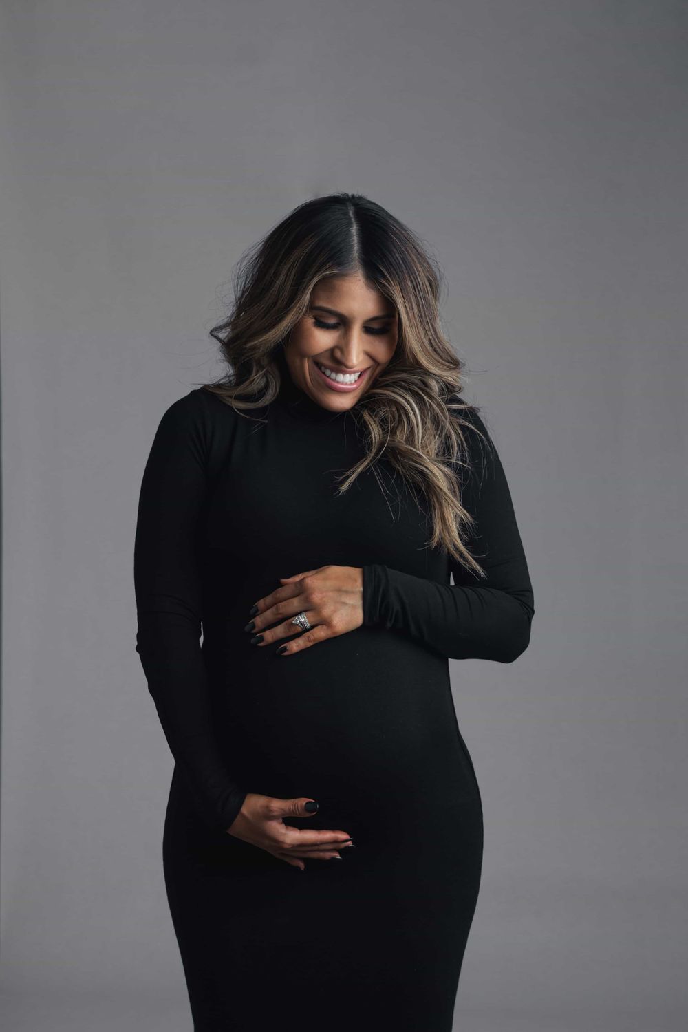 Smiling pregnant woman in black turtleneck maternity dress lovingly holds her baby bump in studio.