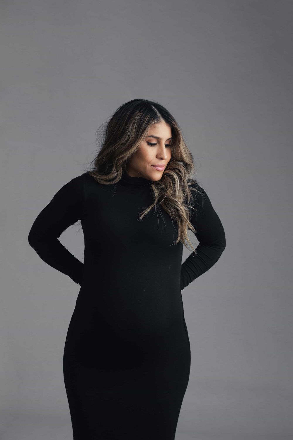 Pregnant woman in elegant black long-sleeve dress poses with hands on hips in professional studio.