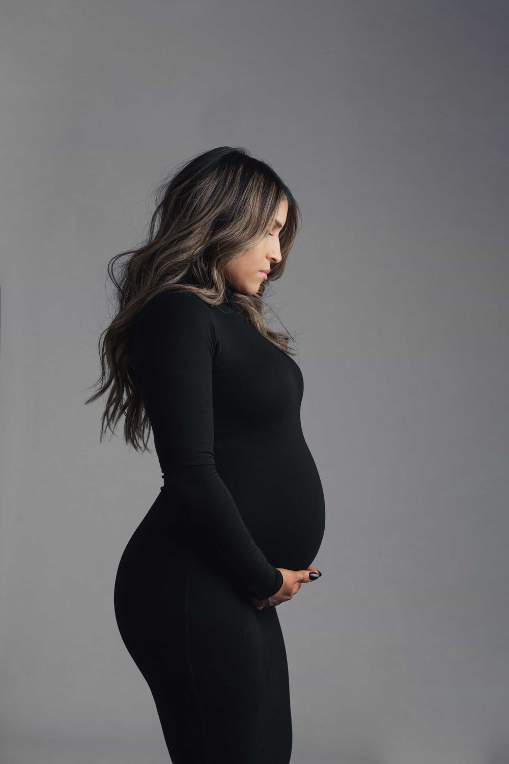 Side profile of pregnant woman in black fitted dress gently holding her baby bump in studio.