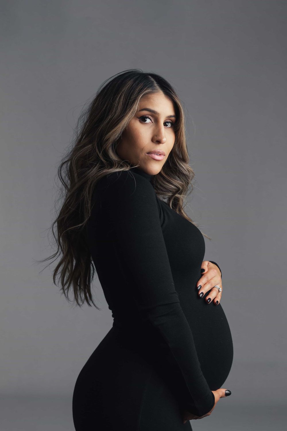 Pregnant woman in black fitted dress poses sideways cradling bump while looking over her shoulder.