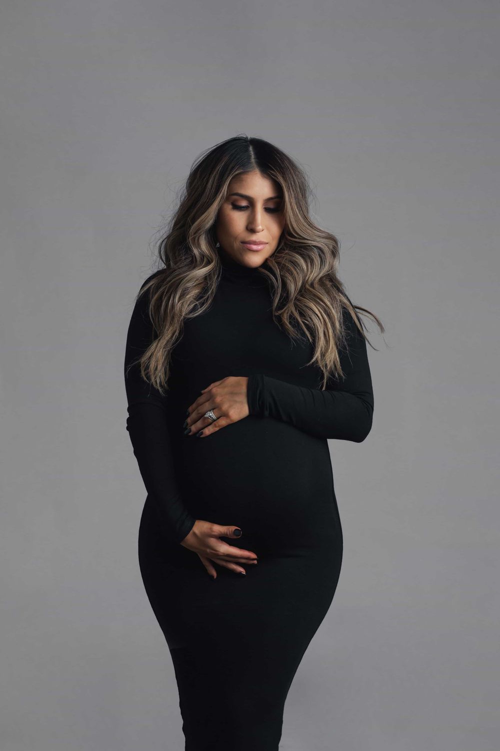 Pregnant woman in black long-sleeve turtleneck dress gently holds baby bump looking down in studio.