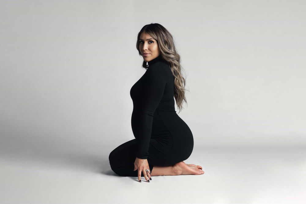 Pregnant woman in black turtleneck outfit sitting sideways on white studio floor.