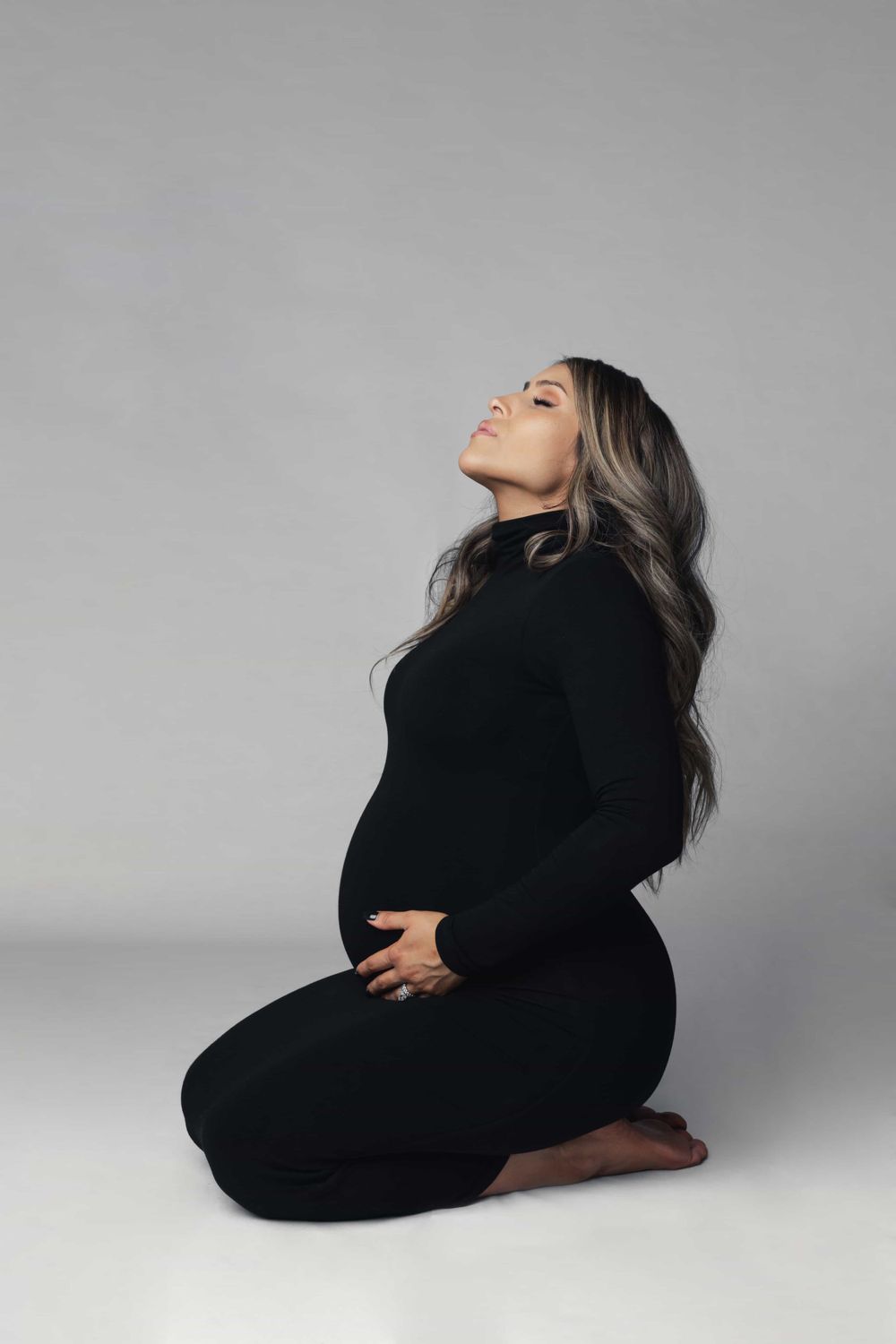 Pregnant woman in black turtleneck dress kneeling with head back in studio maternity shoot.