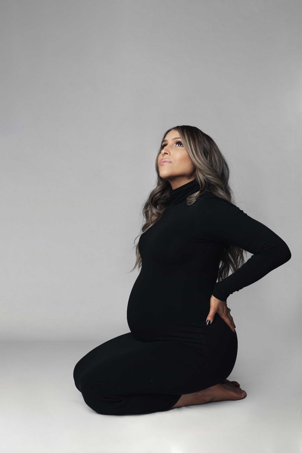 Pregnant woman in black outfit kneeling and looking up in professional studio photo.