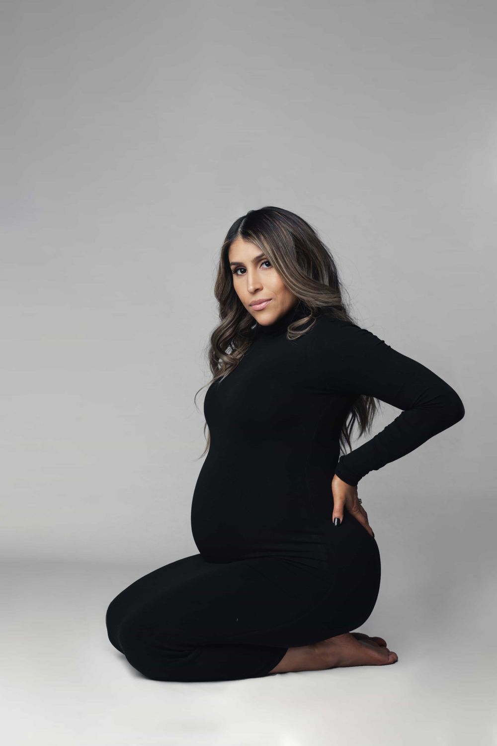 Pregnant woman in black outfit kneeling with hand on back in studio maternity photo.