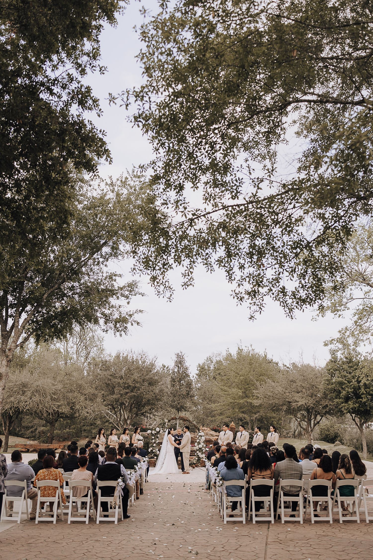 An outdoor wedding ceremony with white chairs arranged in rows under tree canopies.