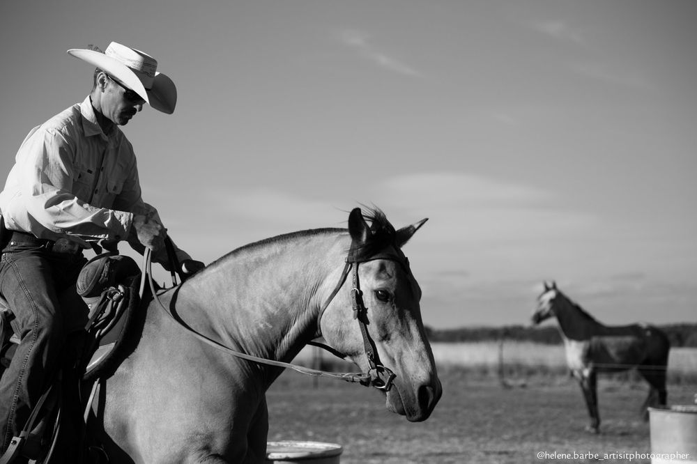 A cowboy rides a light-colored horse in black and white with another horse visible in the background.