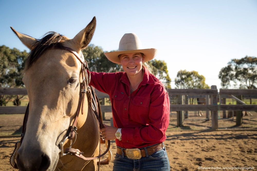 A beautiful dun horse stands with its rider on a sunny ranch with wooden fencing in the background.