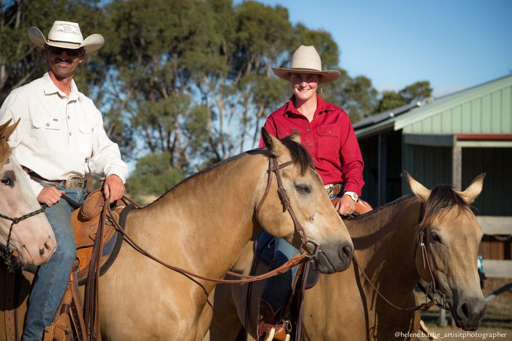 Two horses and their riders pause together near a ranch building on a sunny afternoon.