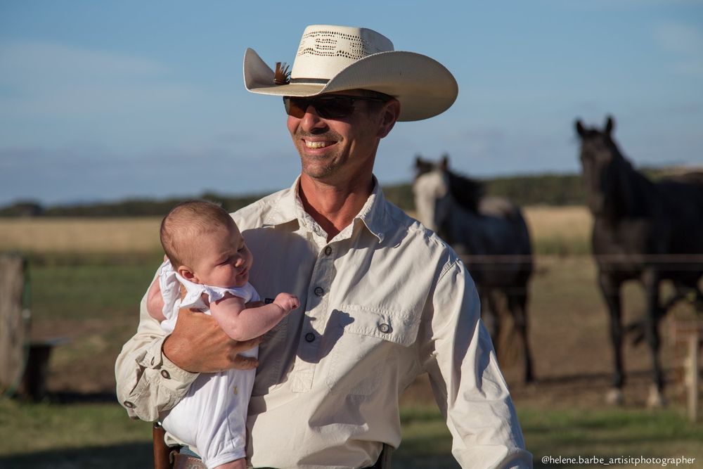 Person in cowboy hat holding a baby outdoors with horses visible in the background.