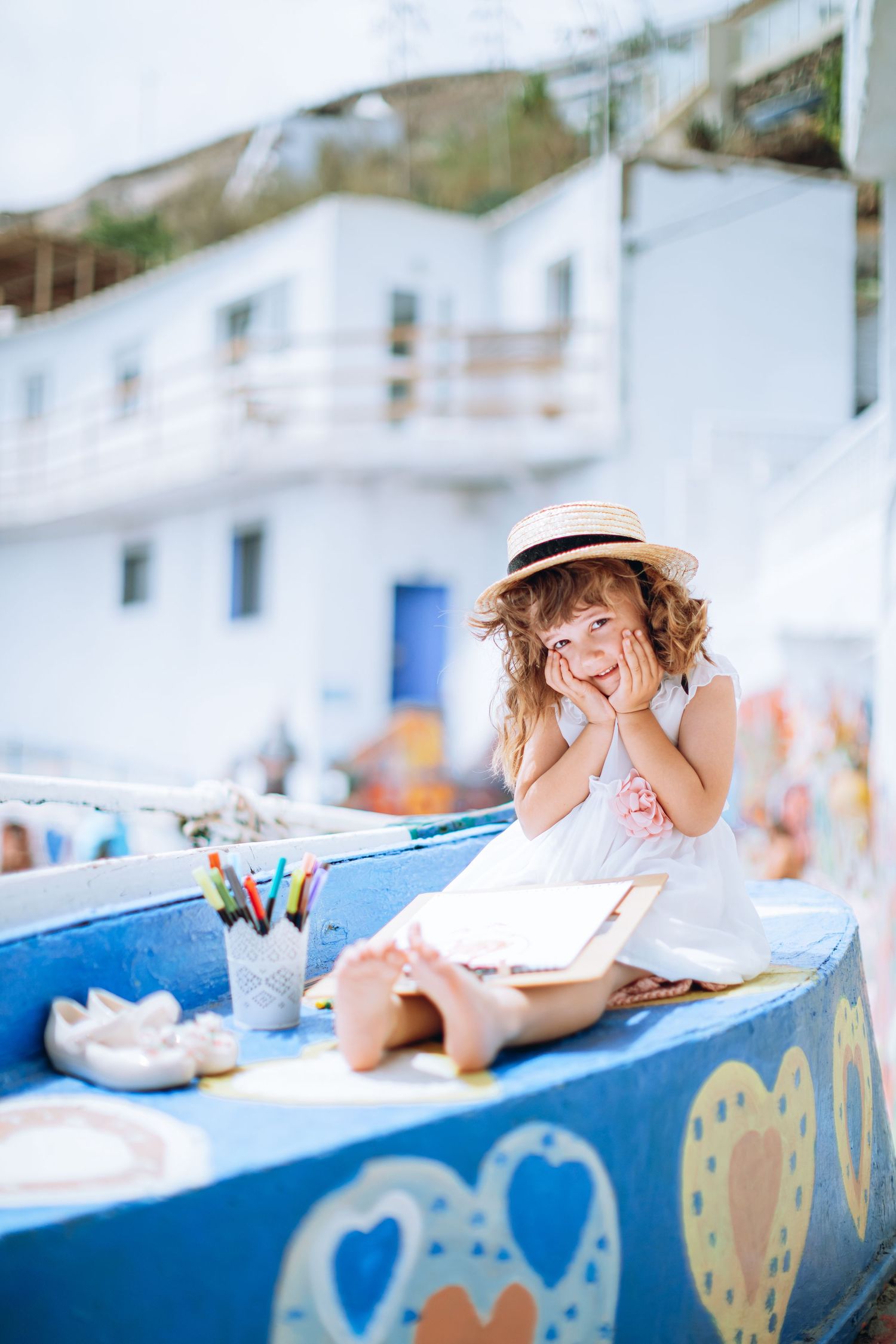 A young girl in a white dress and straw hat smiling while sitting on a blue boat with drawing supplies beside her.