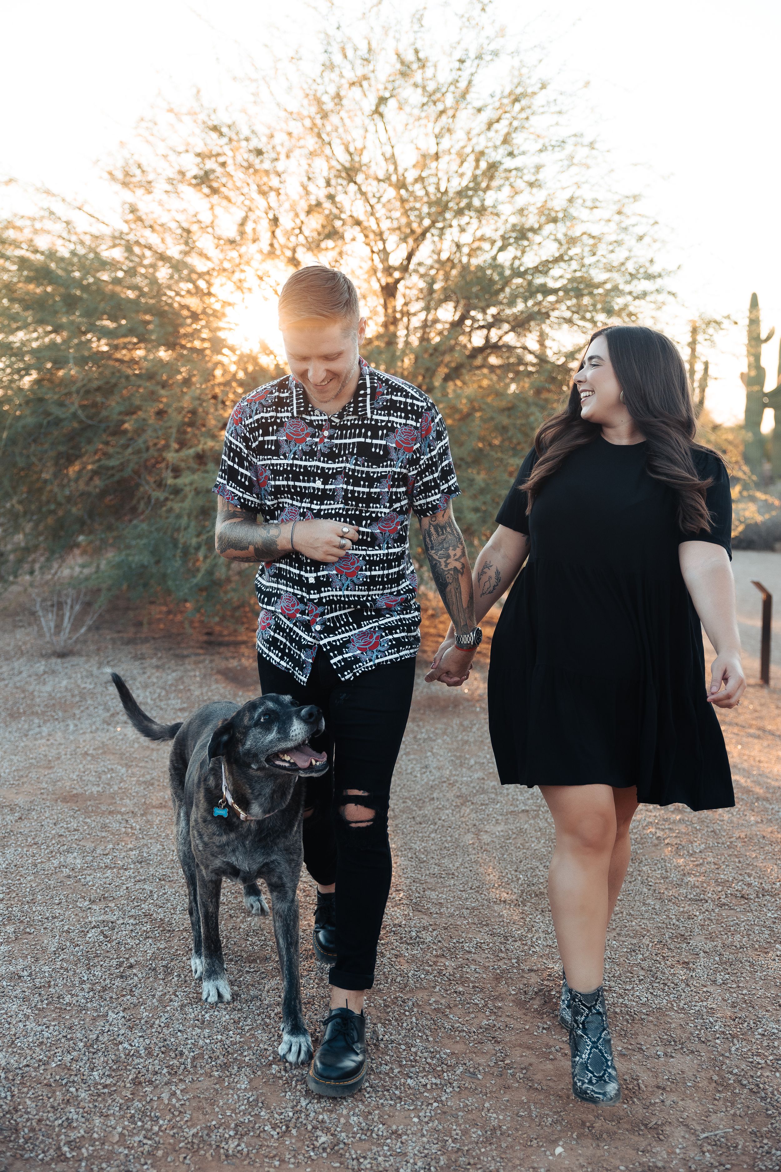 Couple walking their black dog on a desert trail during golden hour with desert plants in background.