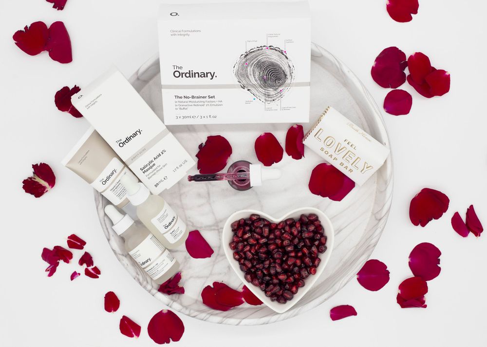 Beauty products arranged with red rose petals and heart shape on white tray.
