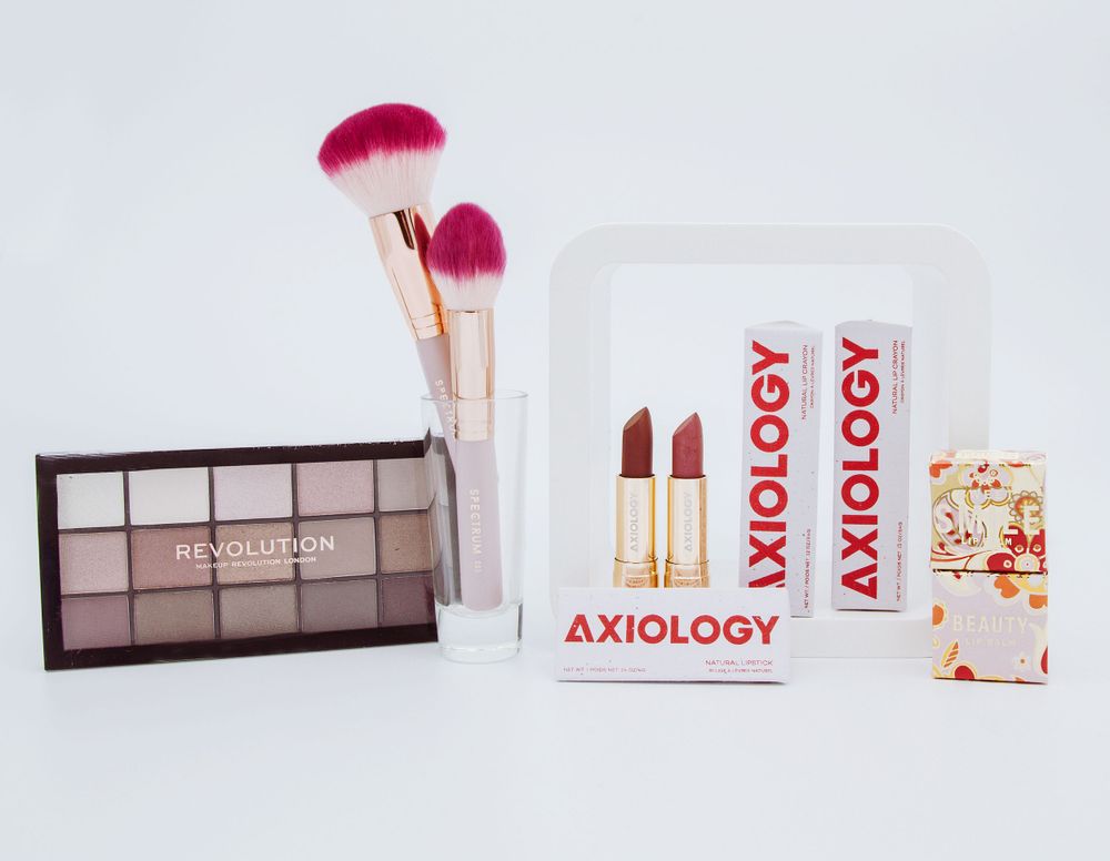 Axiology beauty products including lipsticks, makeup brushes and eyeshadow palette on white background.