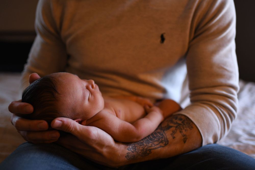 A newborn baby rests peacefully in gentle hands with a tattooed forearm visible.