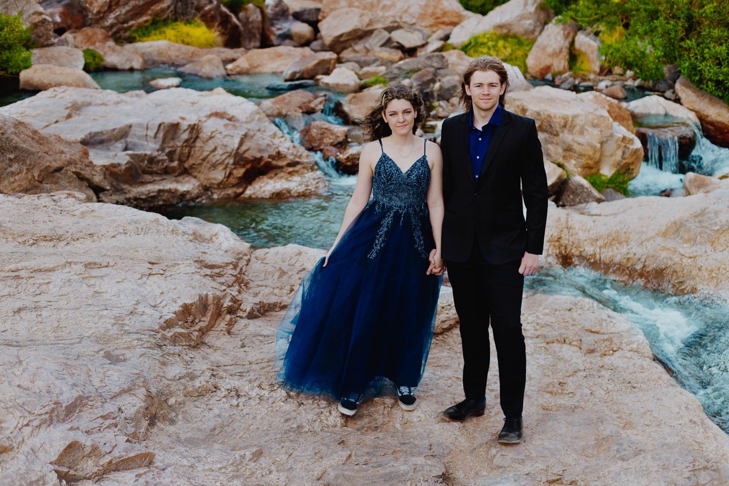 Formal couple poses by flowing mountain stream with blue tulle gown against rocky landscape.