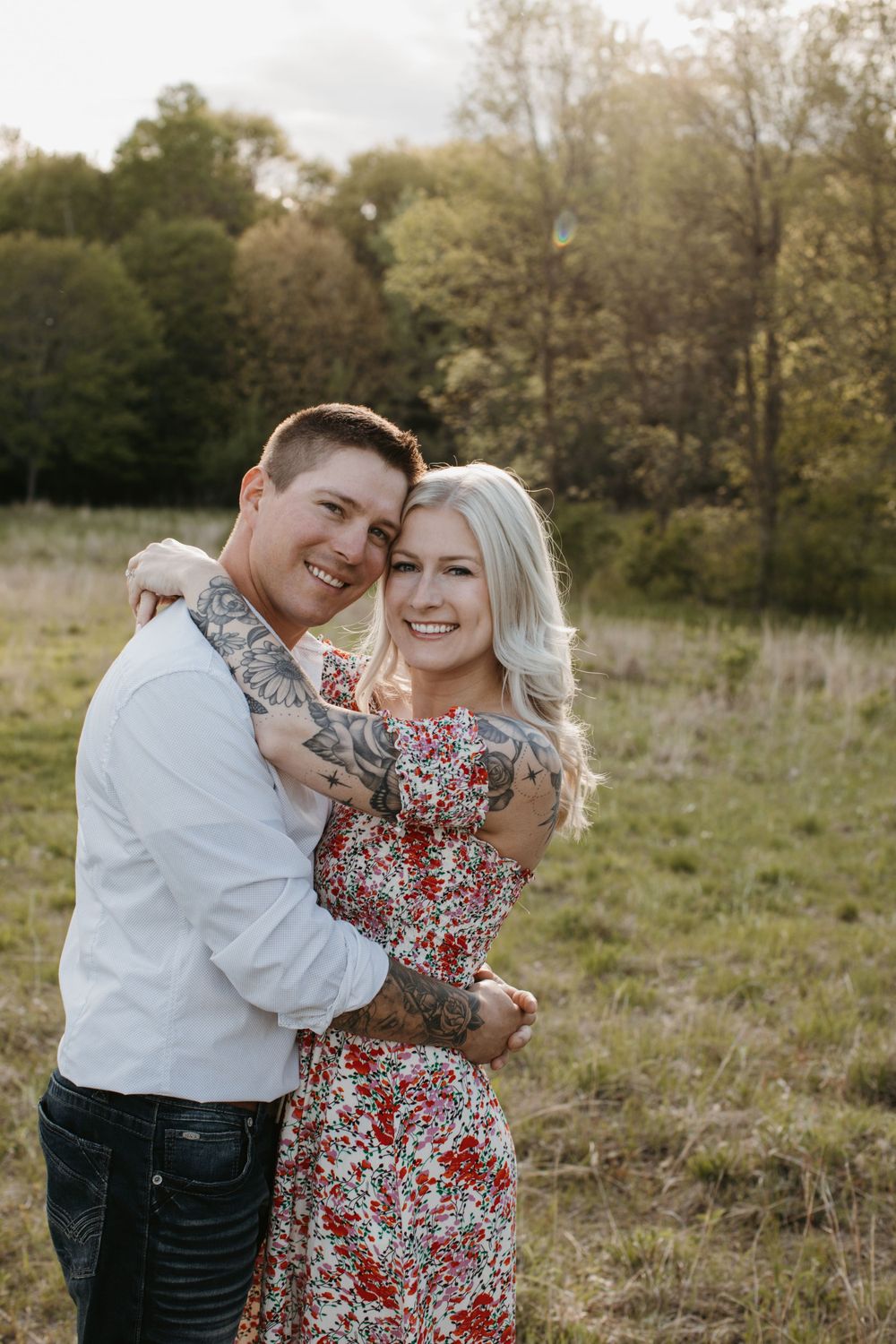 Smiling tattooed couple hugs closely in a golden-lit open field during engagement session.