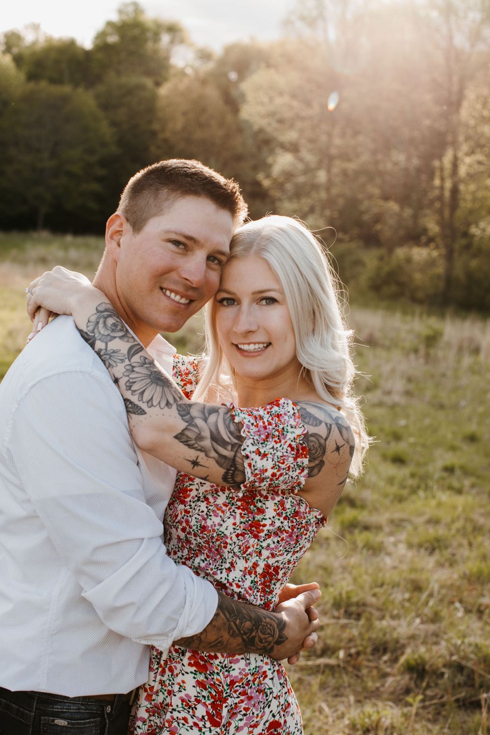 Tattooed couple embraces and smiles in a sunlit meadow during golden hour session.
