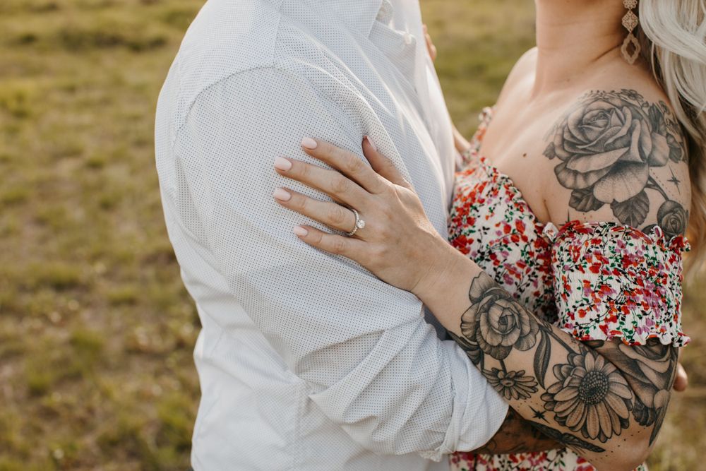 Close-up of tattooed woman embracing partner showing engagement ring in outdoor setting.