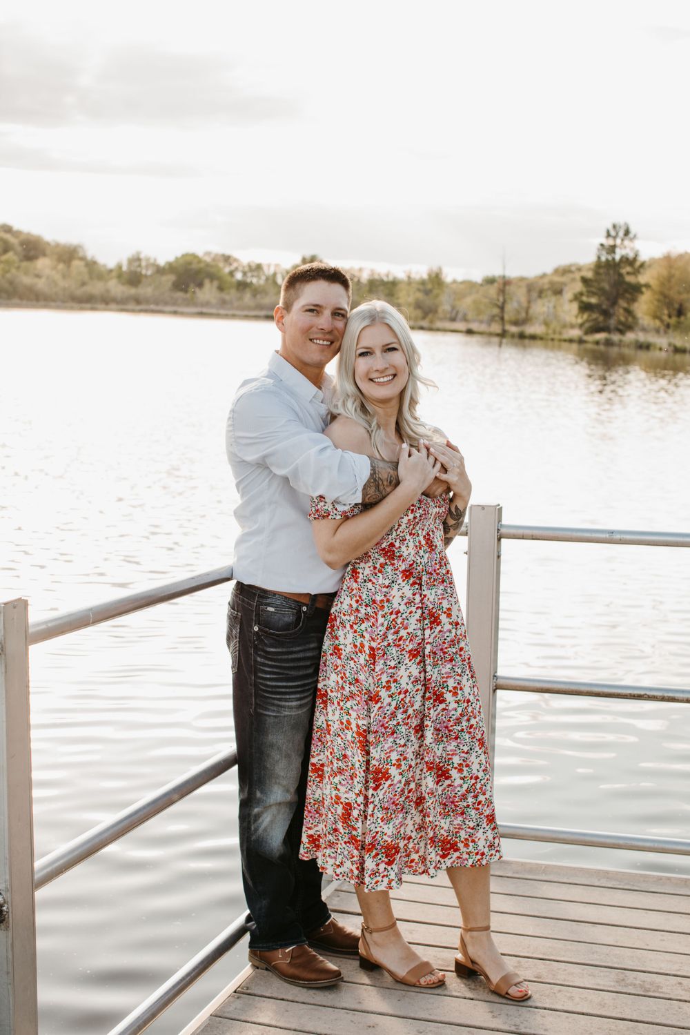 Happy couple embracing on a wooden dock by a calm lake, woman in floral dress and man in white shirt.