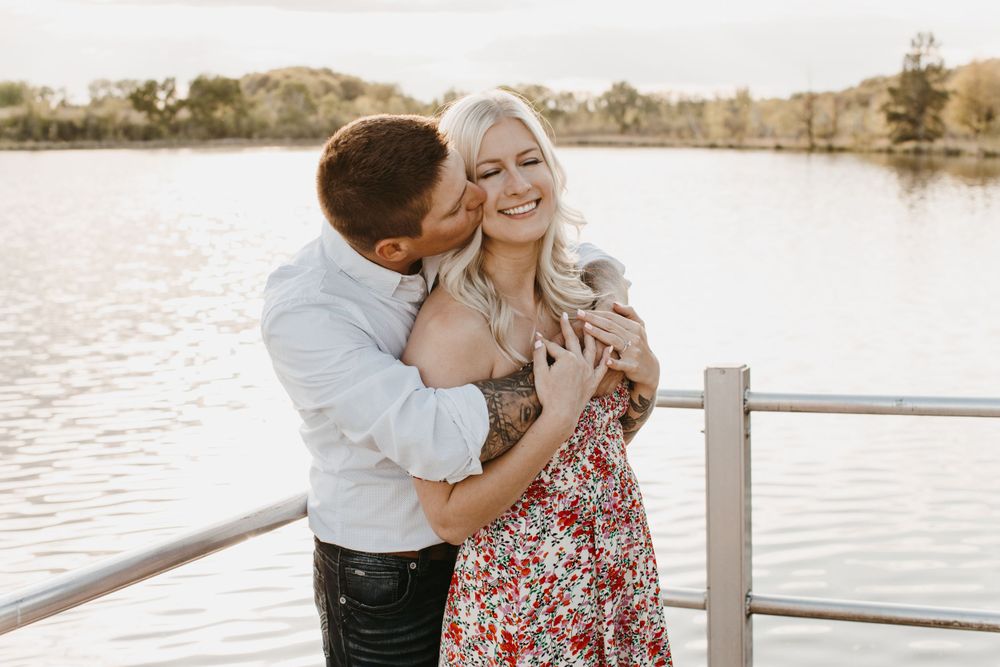 Man kissing smiling woman on cheek on lakeside dock, she wears a red floral dress with tattoos.