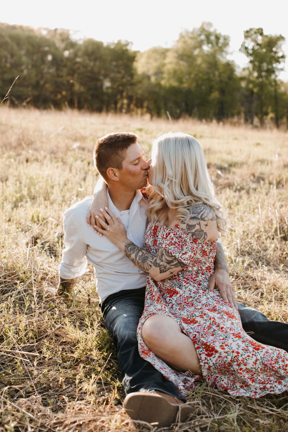 Tattooed couple sharing a kiss while sitting in a golden sunlit field during sunset.