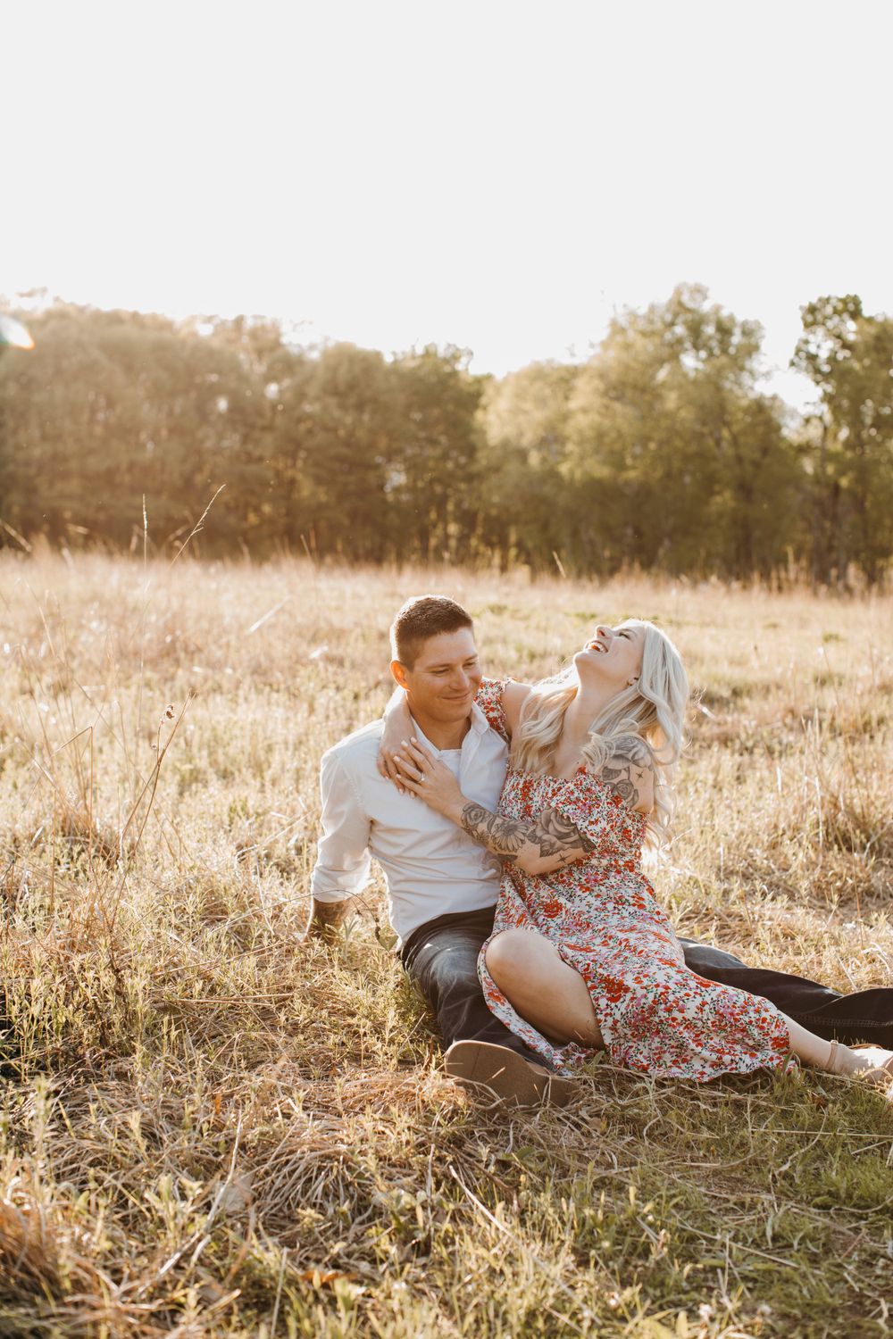 Couple laughing and cuddling in a sunlit meadow, woman in floral dress with tattoos on her arms.