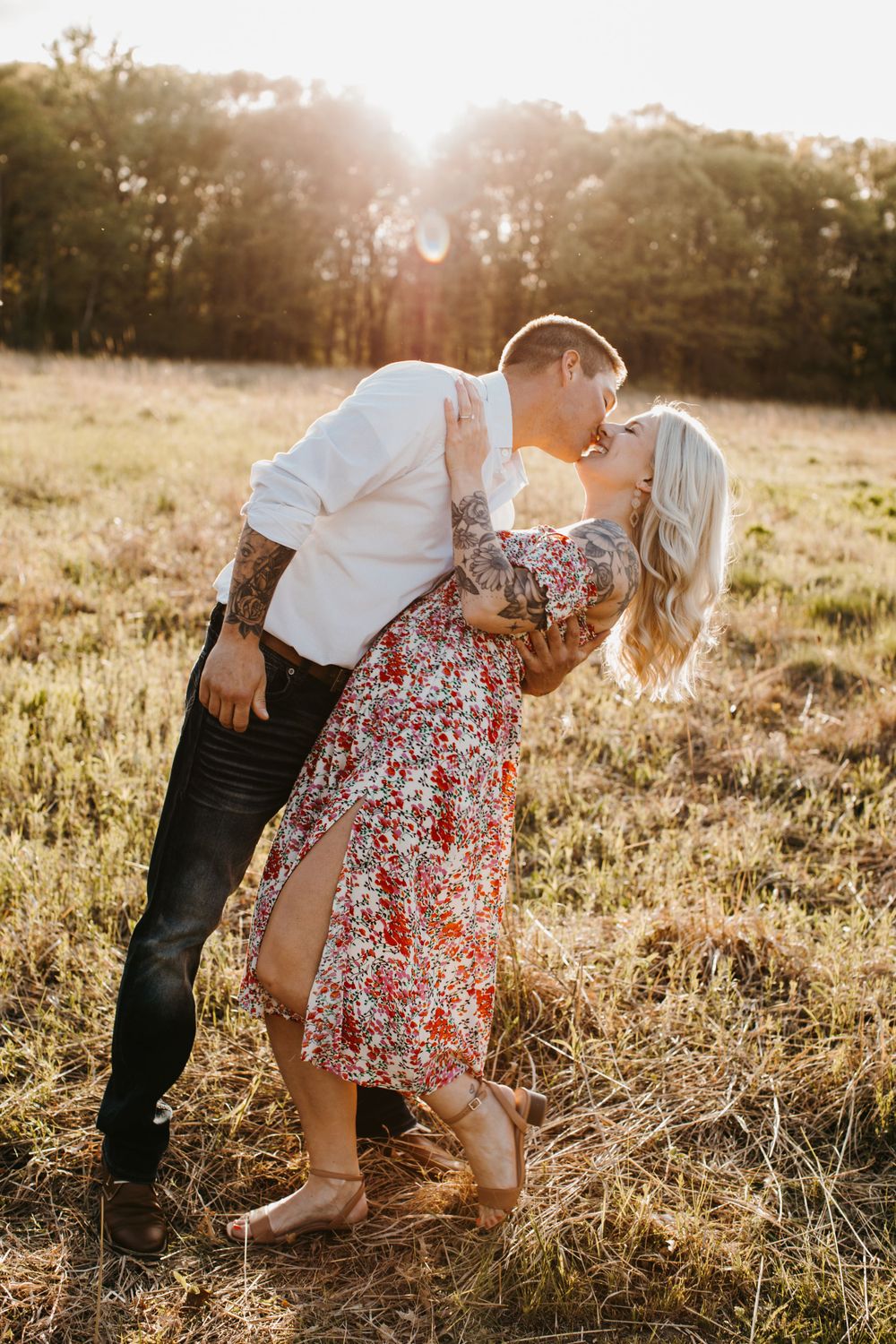 Couple sharing a romantic dip kiss in a golden sunset field, both with visible arm tattoos.