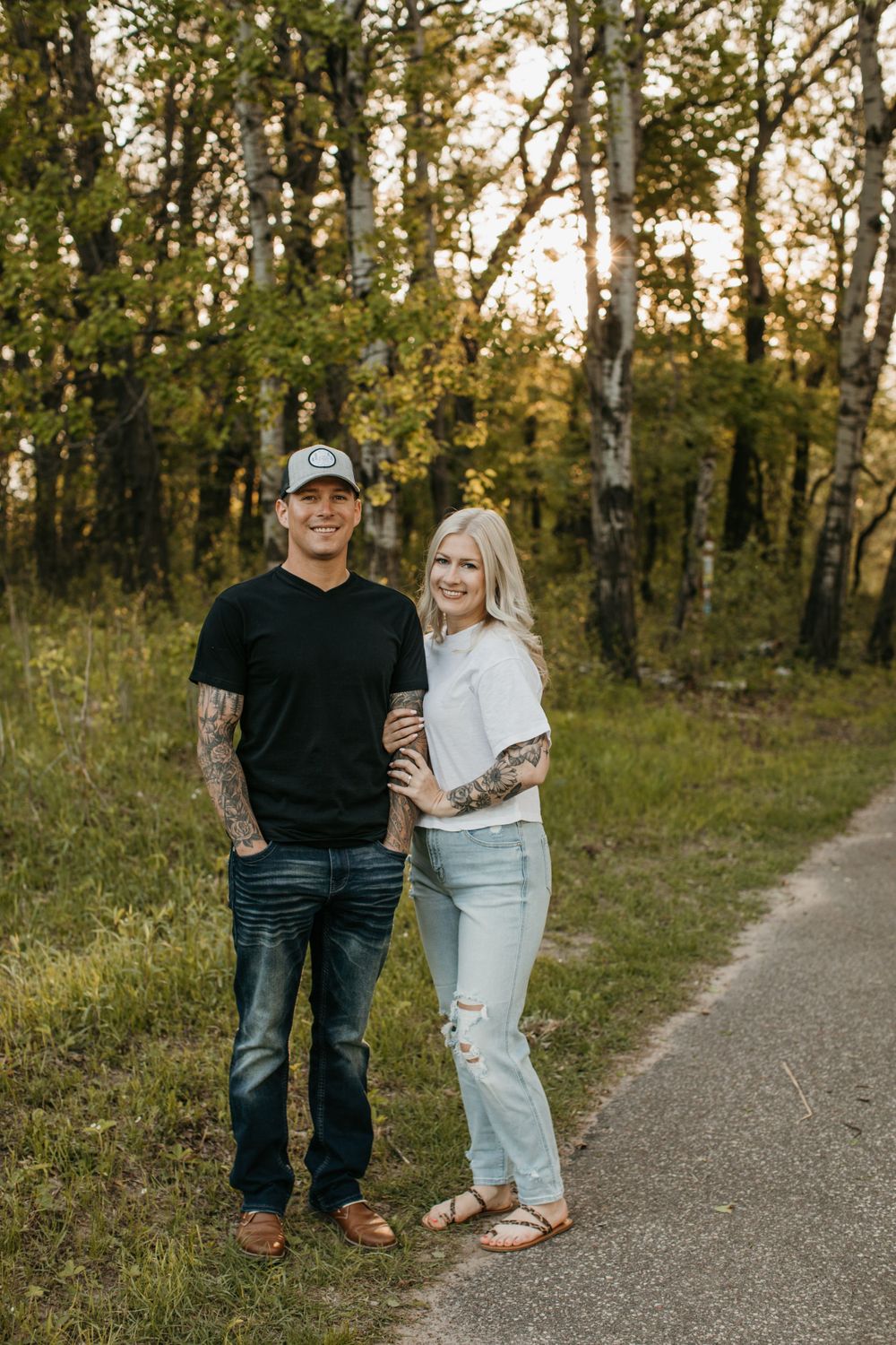 Tattooed couple smiling together on a forest path at sunset, wearing casual black and white outfits.