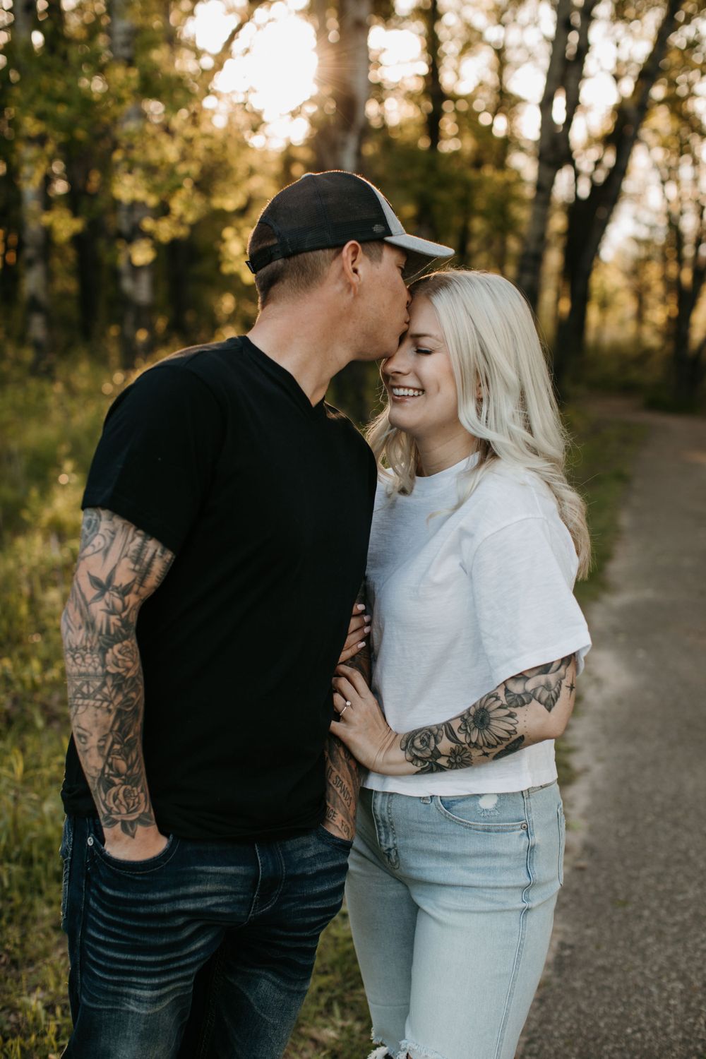 Tattooed man in black shirt kissing woman on forehead on a sunlit forest trail.
