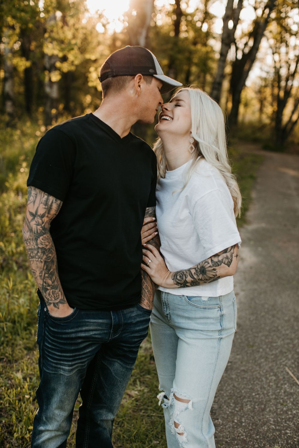 Laughing tattooed couple almost kissing on a tree-lined path in warm golden sunset light.