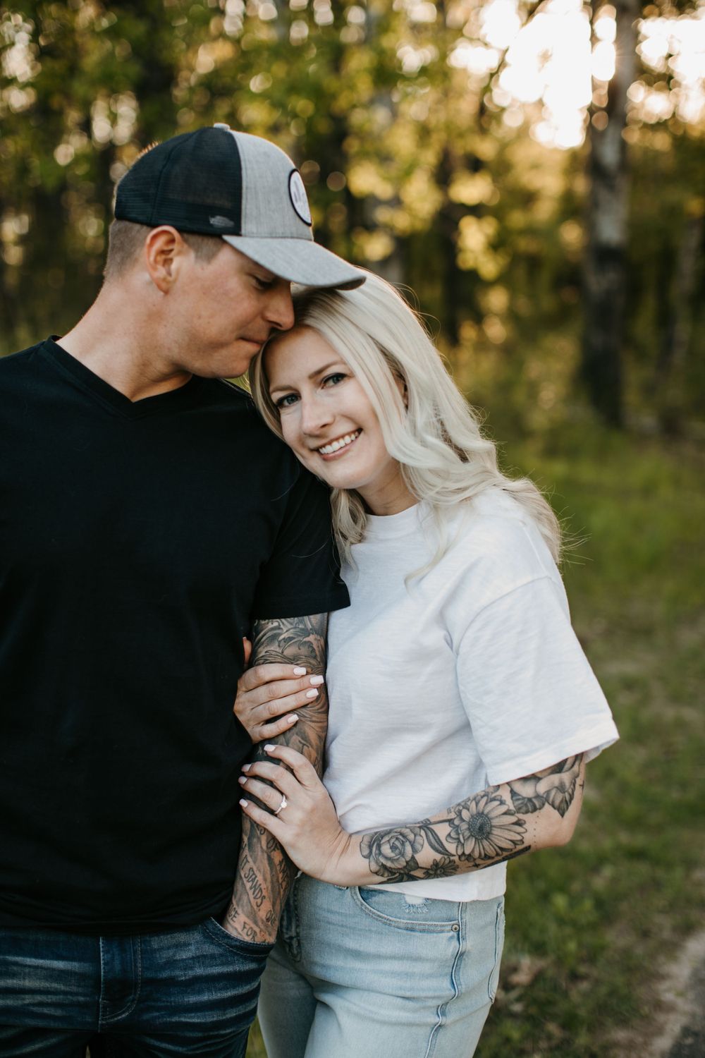 Couple embracing in forest, tattooed woman in white tee smiling, man in black shirt and cap kissing her head.