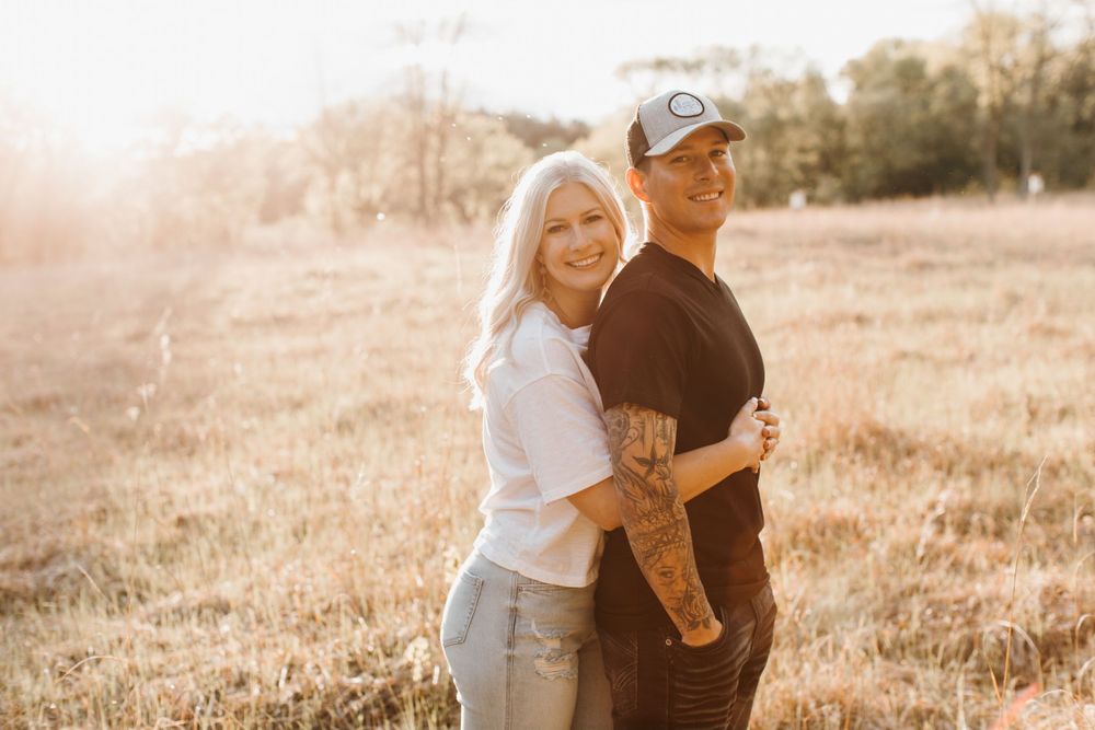 Smiling couple hugging in sunny golden field, woman in white tee and jeans, man in black shirt and cap.