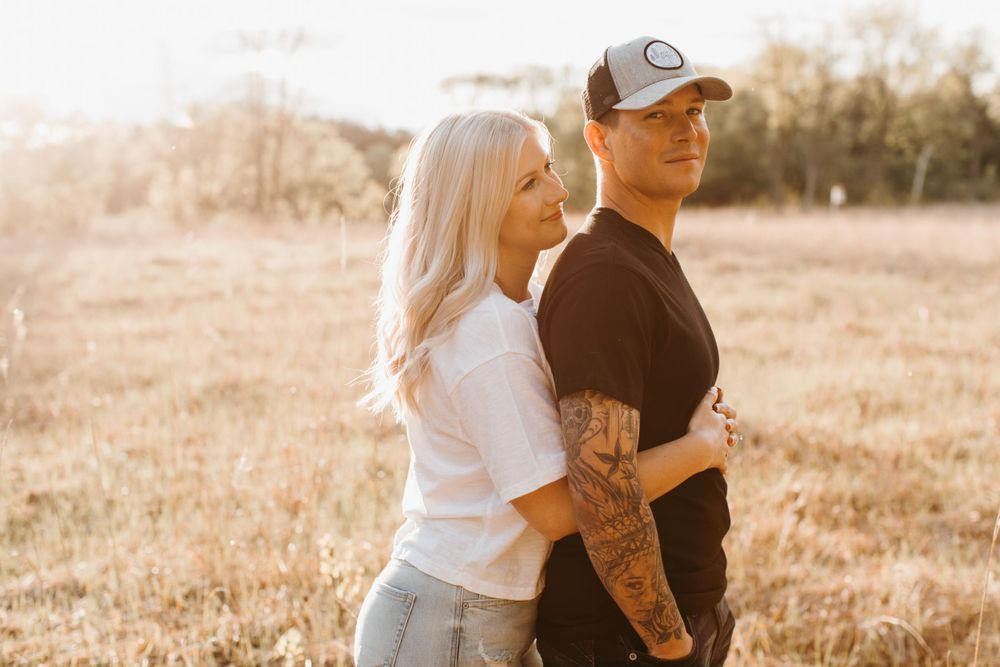 Tattooed couple standing back-to-back in sunlit field, woman in white tee, man in black shirt and cap.