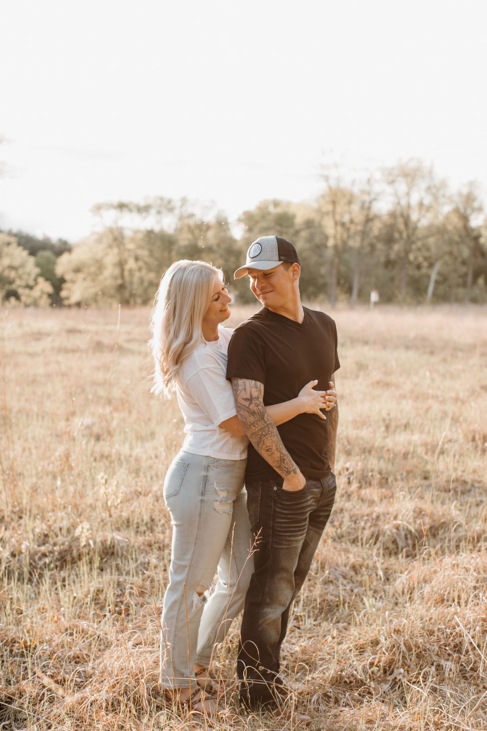 Tattooed couple laughing together in sunny dry grass field, woman in white tee and jeans, man in cap.
