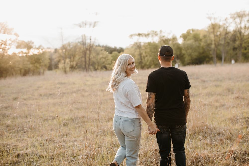 Couple holding hands walking away through golden field, woman in white tee and jeans, man in black shirt.