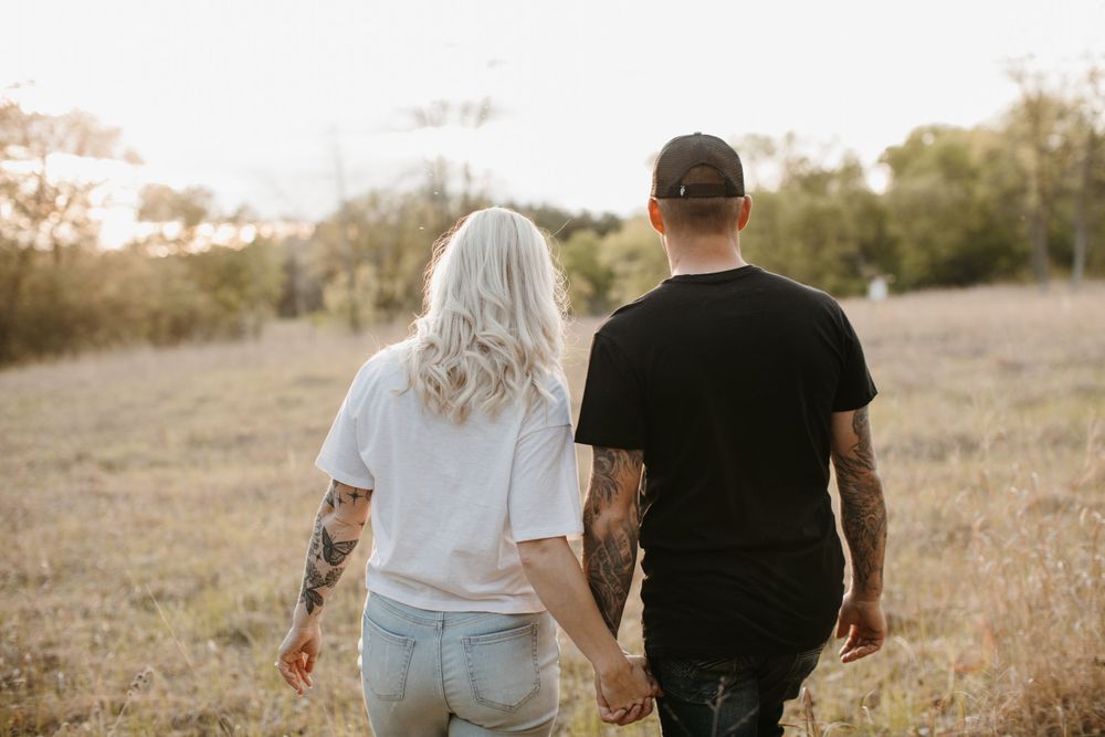 Tattooed couple holding hands walking through golden field, viewed from behind, casual outdoor setting.