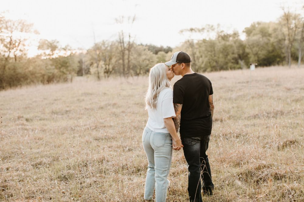 Couple sharing a kiss while holding hands in a sunny open field, woman in white tee and light jeans.