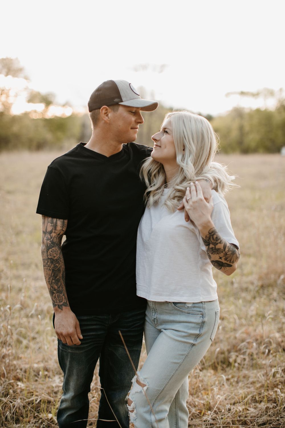 Tattooed couple gazing at each other in golden field, man in black shirt and cap, woman in white tee.